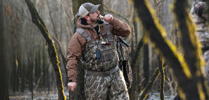 A man standing in the timber blowing a duck call. He is wearing a pair of Gator Waders in Mossy Oak Bottomland. 