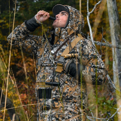 A guy standing in the timber blowing on a duck call while hunting.