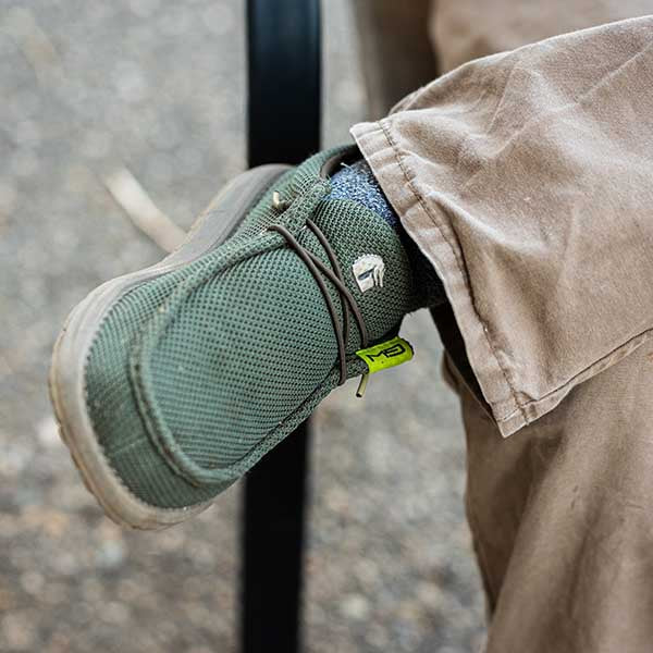 A guy crossing his legs and wearing a pair of Camp Shoes. The color of the shoes is olive.