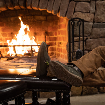 A guy propping his feet up by a fireplace while wearing a pair of olive Camp Shoes.