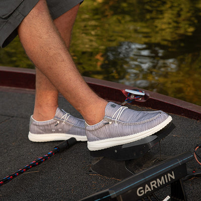 A guy on a boat with his foot on a trolling motor with Camp Shoes on. The shoes are grey.