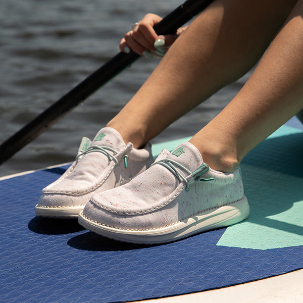 A girl sitting on a paddle board in the water wearing Camp Shoes. The shoes are a white color with teal accents.