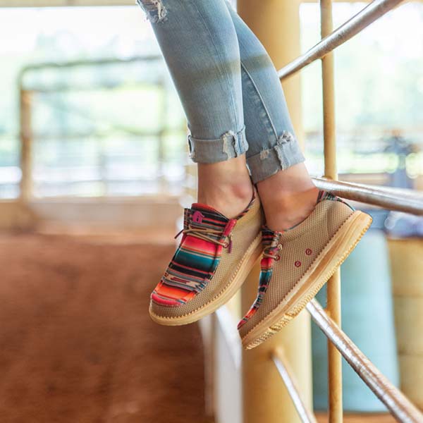 A girl sitting on a gate with her feet dangling and wearing a pair of serape Camp Shoes.
