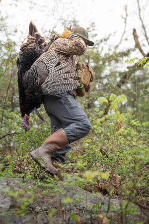 A guy carrying a turkey over his shoulder while walking on rocks in a pair of Gator Waders Omega Flow boots in the color brown.