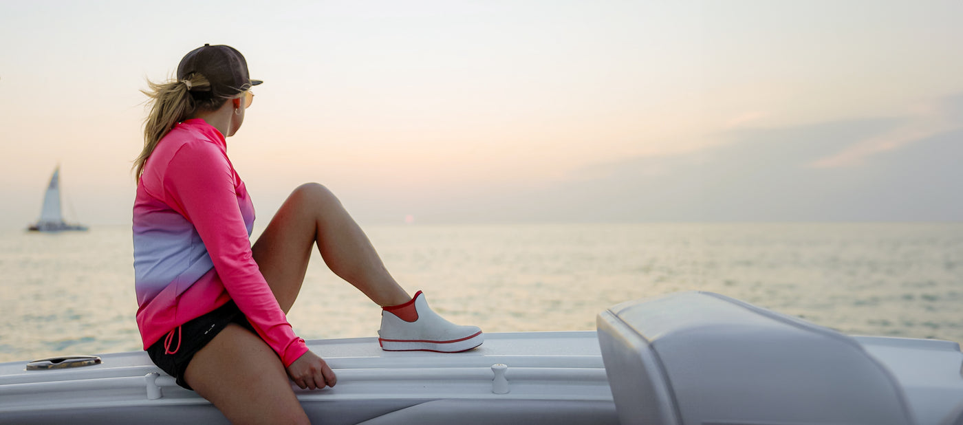 woman sitting on the side of a boat looking out at the lake while wearing a pair of Gator Wader's Air Mesh Camp Boots in the style coral